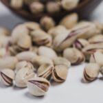 Beyaz bir zemin üzerinde, yakın plandan çekilmiş, kabuklu kavrulmuş Antep fıstıklarının dağınık görüntüsü. Odak noktası, ön plandaki bir fıstık tanesi üzerindedir.