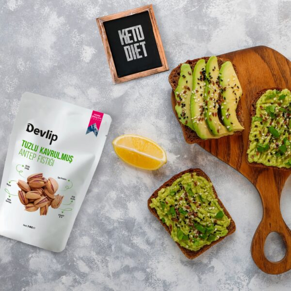 Gri bir zemin üzerinde, "Keto Diet" yazılı küçük bir levha ve üzerinde avokado dilimleri olan iki dilim ekmek bulunuyor. Yanında Devlip Tuzlu Kavrulmuş Antep Fıstığı paketi, limon dilimi ve bir kaşık yer alıyor.