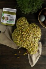Ahşap bir masa üzerinde, bir bezin üzerinde Antep Fıstığı Kreması sürülmüş ve bolca fıstık parçacıklarıyla süslenmiş yonca şeklinde bir pasta duruyor. Pastanın yanında Devlip Antep Fıstığı Kreması kavanozu ve bir sepet içinde renkli yumurtalar görünüyor.